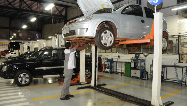 Mecânico conferindo a parte inferior do motor de carro cinza suspenso no elevador da oficina da concessionária; É preciso ficar atento às sugestões de manutenção do carro em concessionárias e oficinas, pois muitas vezes tentam empurrar serviços desnecessários