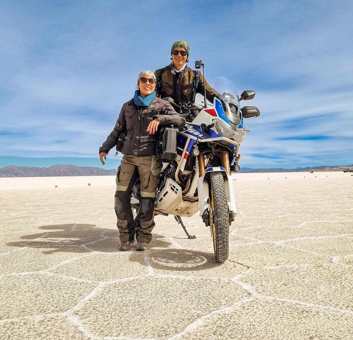 Casal equipado com roupas de proteção para viagens de moto sobre motocicleta com fundo de céu azul em deserto de sal