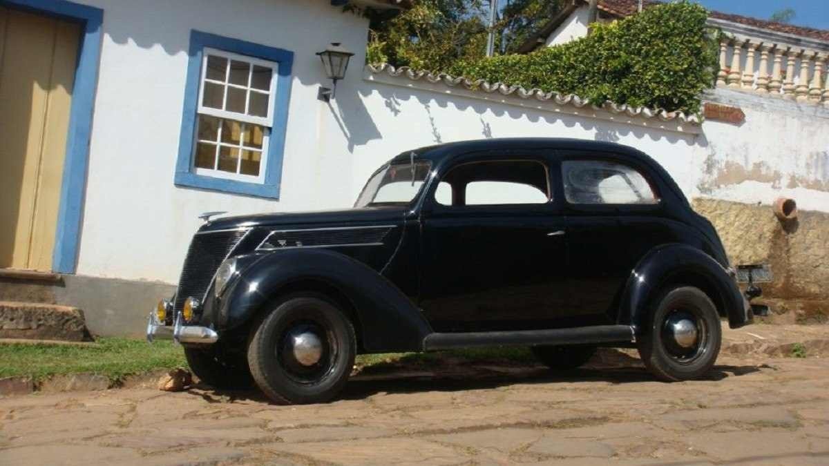 Carro antigo preto está estacionado em rua de pedra, ao fundo há uma casa de época branca com entorno da porta e janela em azul claro.