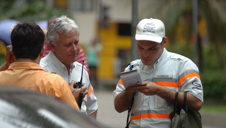 Guarda municipal de Belo Horizonte com vestes brancas e listras laranjas neon conversa com homem branco de cabelos grisalhos ao seu lado. Pessoa com cabelo preto e blusa social laranja é vista no canto esquerdo da foto