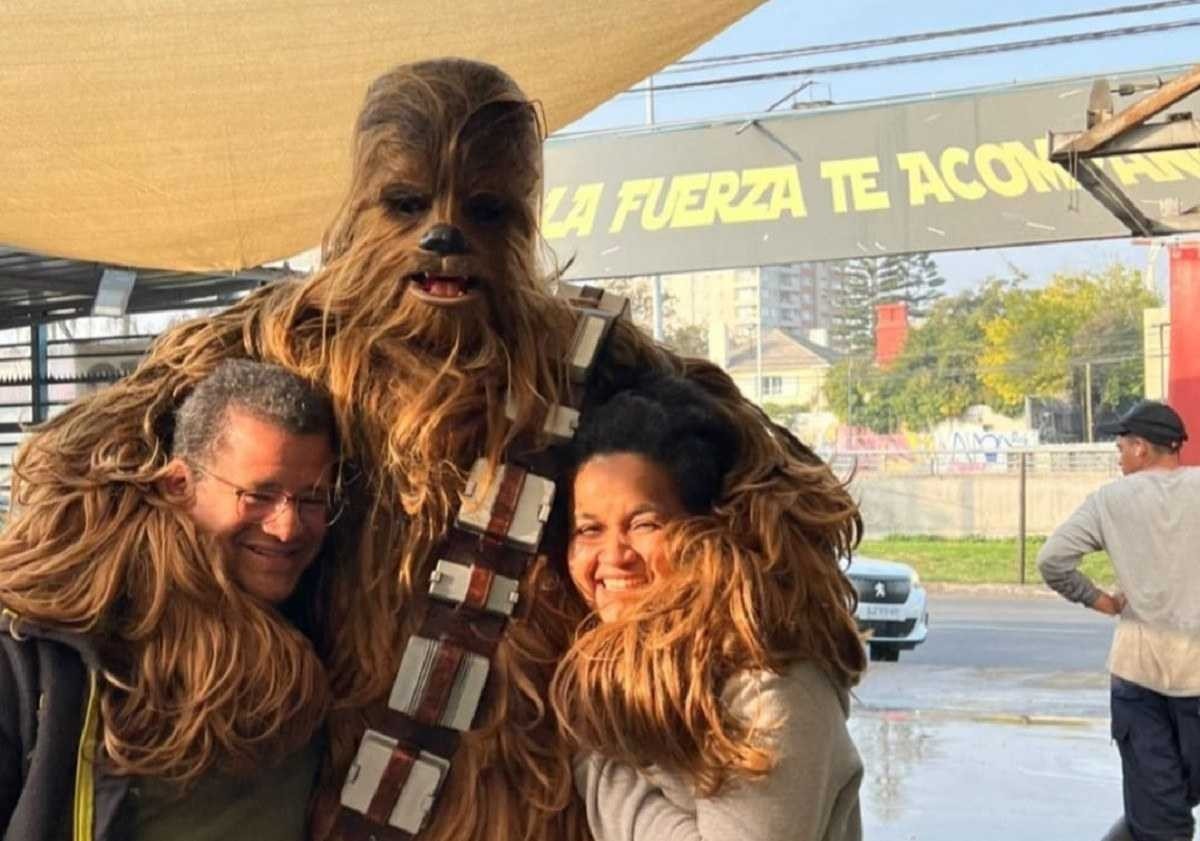 Chewbacca está abraçando duas pessoas, ao fundo é possível ver uma placa de lava jato.