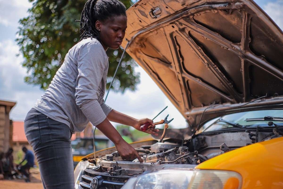 Mulher negra com moletom cinza e calças jeans conserta um carro amarelo.