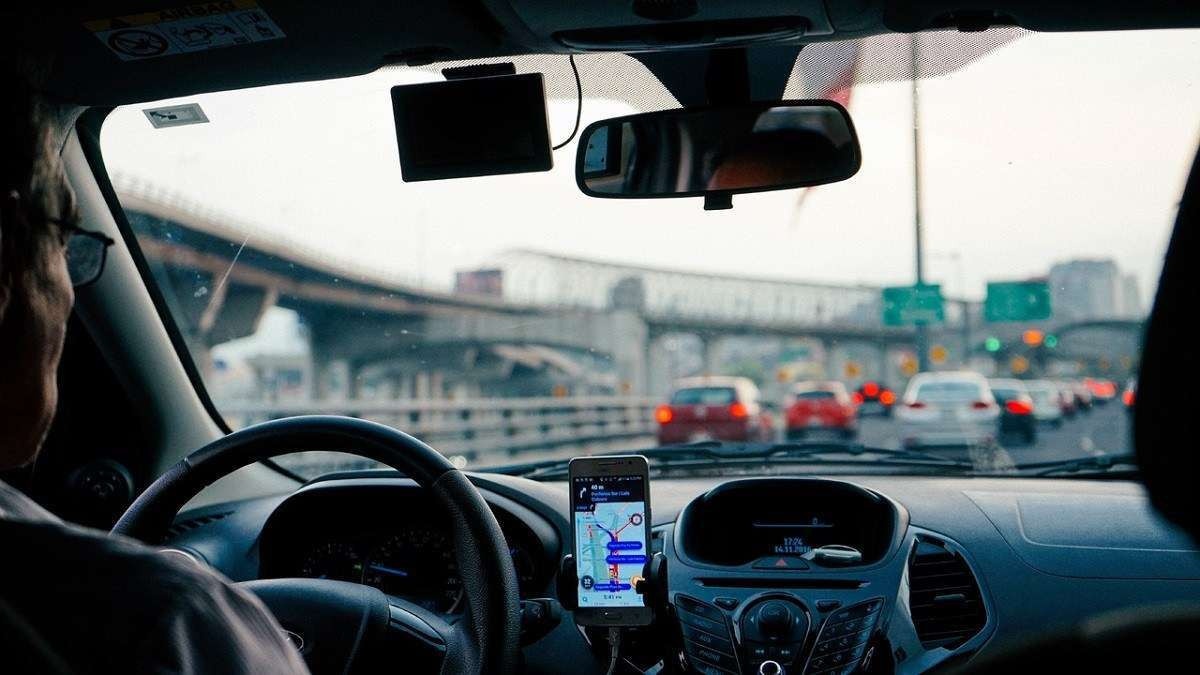 Pessoa dirige carro enquanto confere o GPS, através do para-brisa é possível ver placas de destino e carros andando.