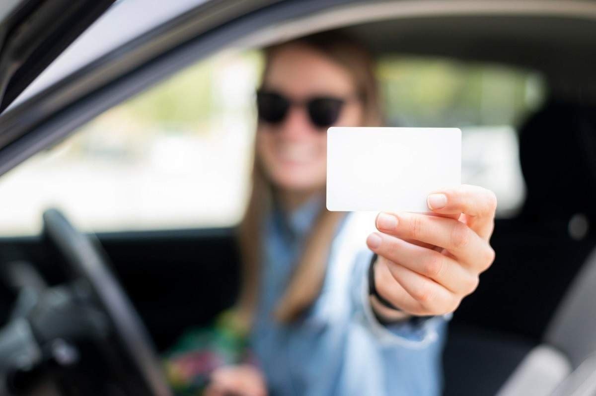 Mulher branca com óculos dentro de um carro mostrando um documento todo branco.