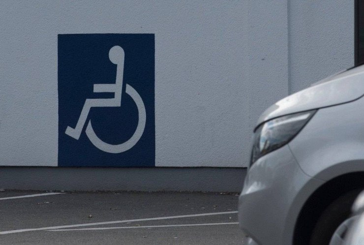 Pequena parte de um carro prata &eacute; visto no canto direito, ao fundo h&aacute; um desenho de uma pessoa em uma cadeira de rodas.