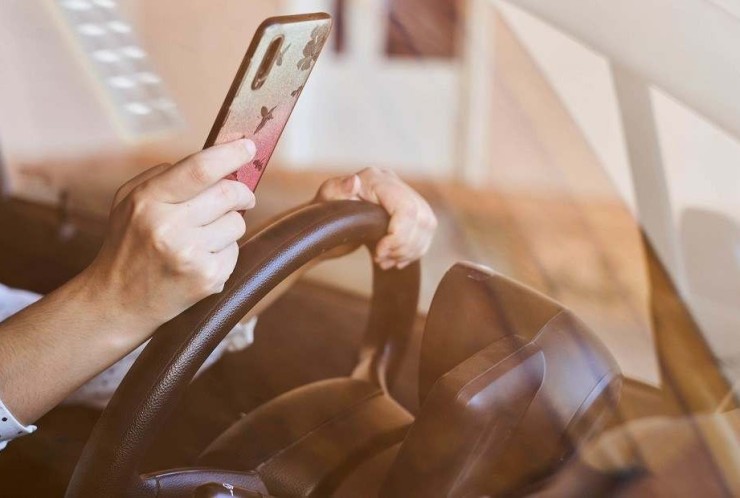 Mulher vestindo blusa de mana longa branca com bolinhas pretas esta com celular na m&atilde;o apoiada no volante.