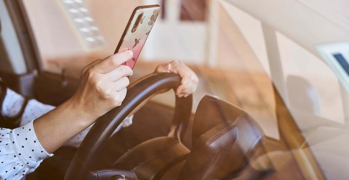 Mulher vestindo blusa de mana longa branca com bolinhas pretas esta com celular na mão apoiada no volante.