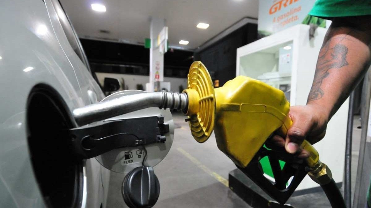 Bico de bomba de gasolina abastecendo o tanque de um carro prata em posto de combustível