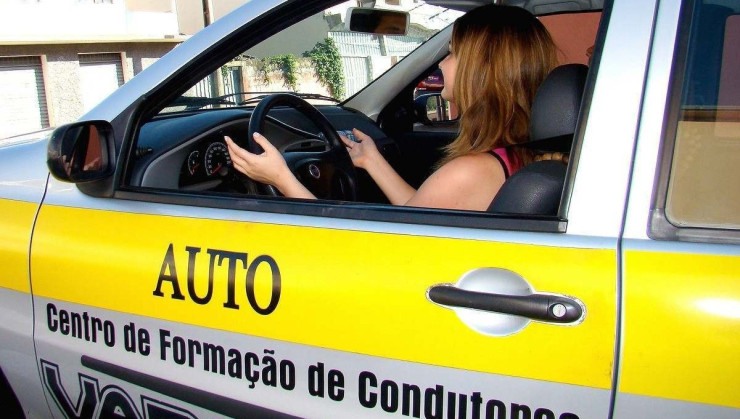 Mulher de cabelo ruivo dirige carro de auto escola, ao fundo é possível ver algumas casas de muro de xapisco e outro coberto por plantas. 
