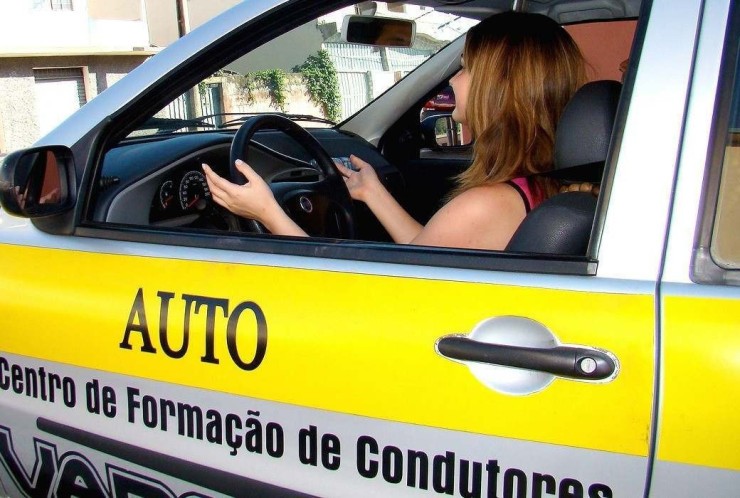 Mulher de cabelo ruivo dirige carro de auto escola, ao fundo é possível ver algumas casas de muro de xapisco e outro coberto por plantas. 