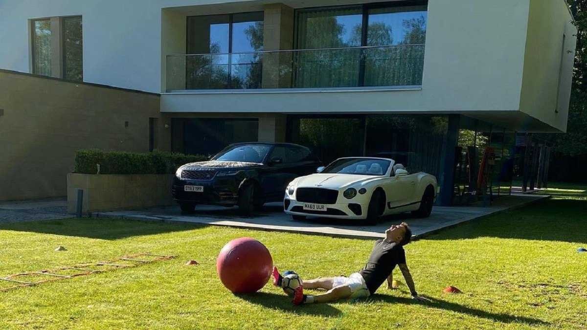 Bernard sentado em grama com Bentley Continental GTC e Range Rover ao fundo.