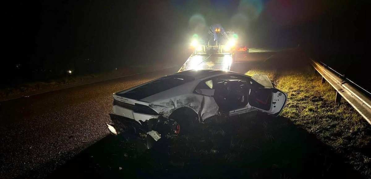 Lamborghini Huracan branca destruída em via pavimentada com veículo com farol ligado ao fundo, barreira de proteção e árvores.