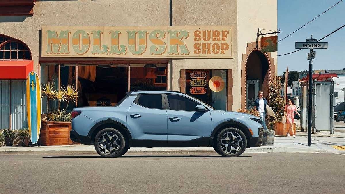 Hyundai Santa Cruz azul estacionada em rua pavimentada com loja de surf ao fundo.