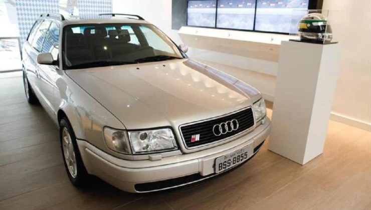 Audi S4 Plus Avant prata dentro de casa com capacete amarelo e verde de Ayrton Senna dentro de um expositor de vidro quadrado.