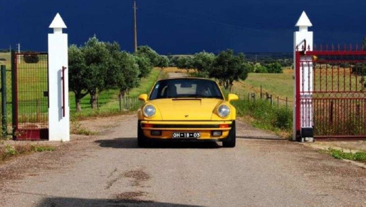 Porsche 911 Carrera Cabriolet amarelo passa por um portão com barras vermelhas e portal branco.