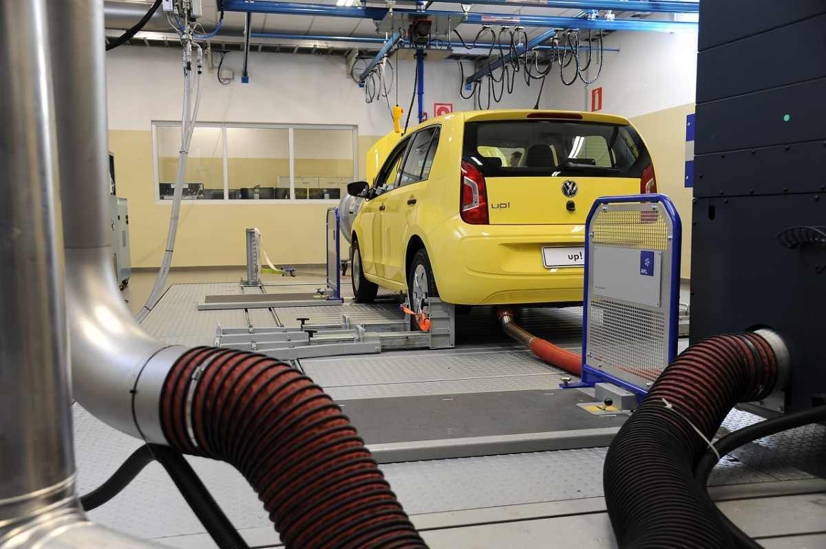 Automóvel up! amarelo, vistpo a partir da traseira, com mangueira acoplada à saída do escapamento,  dentro dos laboratórios de motores e emissões da Volkswagen