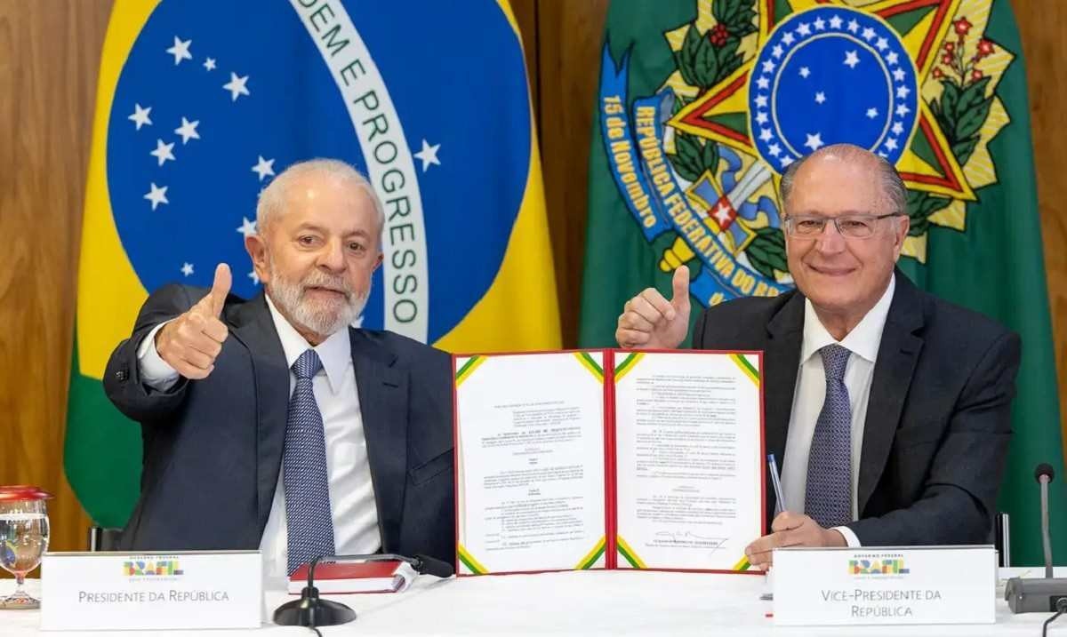 O presidente Luiz Inácio Lula da Silva ao lado do vice-presidente Geraldo Alckmin, durante a cerimônia de Assinatura de Atos relacionados ao Programa Mover: eles estão sentados lado a lado em uma mesa, segurando, em posição central, a pasta com o texto das diretrizes do programa; com a outra mão, ambos fazem sinal de positivo