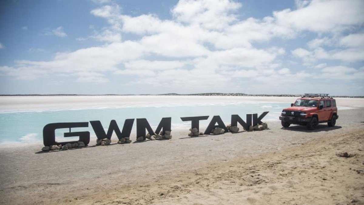 GWM Tank 300 laranja é visto de frente ao lado de placa escrito GW Tank em praia, ao fundo há o mar com água clara e céu azul no topo