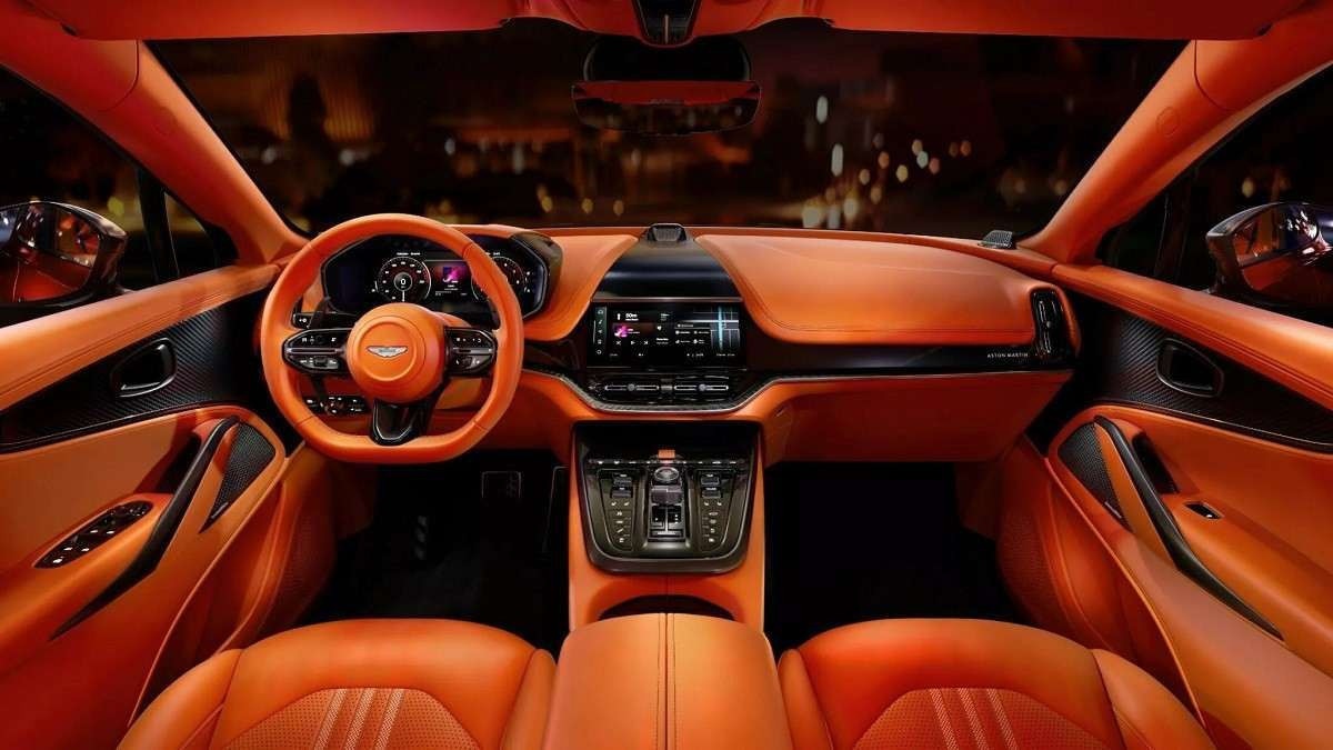 Interior do Aston Martin DBX707 laranja com pequenos detalhes em preto. Pelo para-brisas e janelas laterais é possível ver imagem de cidade distorcida