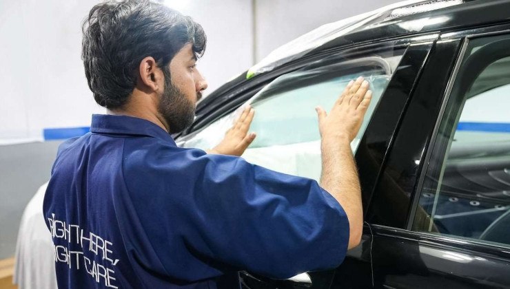 Homem de barba e cabelo curtos e pretos aplica película na janela dianteira esquerda de veículo preto.