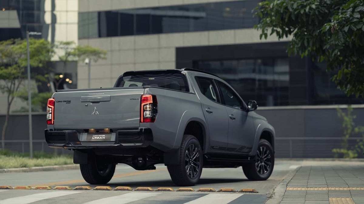 Mitsubishi L200 Triton Sport Urban cinza vista pela traseira está parada em rua pavimentada com grama e pequeno coqueiro do lado esquerdo. Prédio cinza com janelas em preto é visto ao fundo.