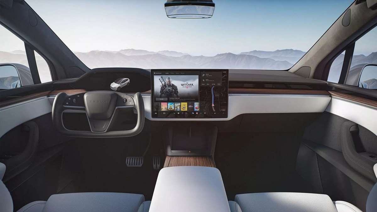 Interior do Tesla Model X com tela de infoentretenimento ligada e céu claro com montanhas visto pelo para-brisas e janelas.
