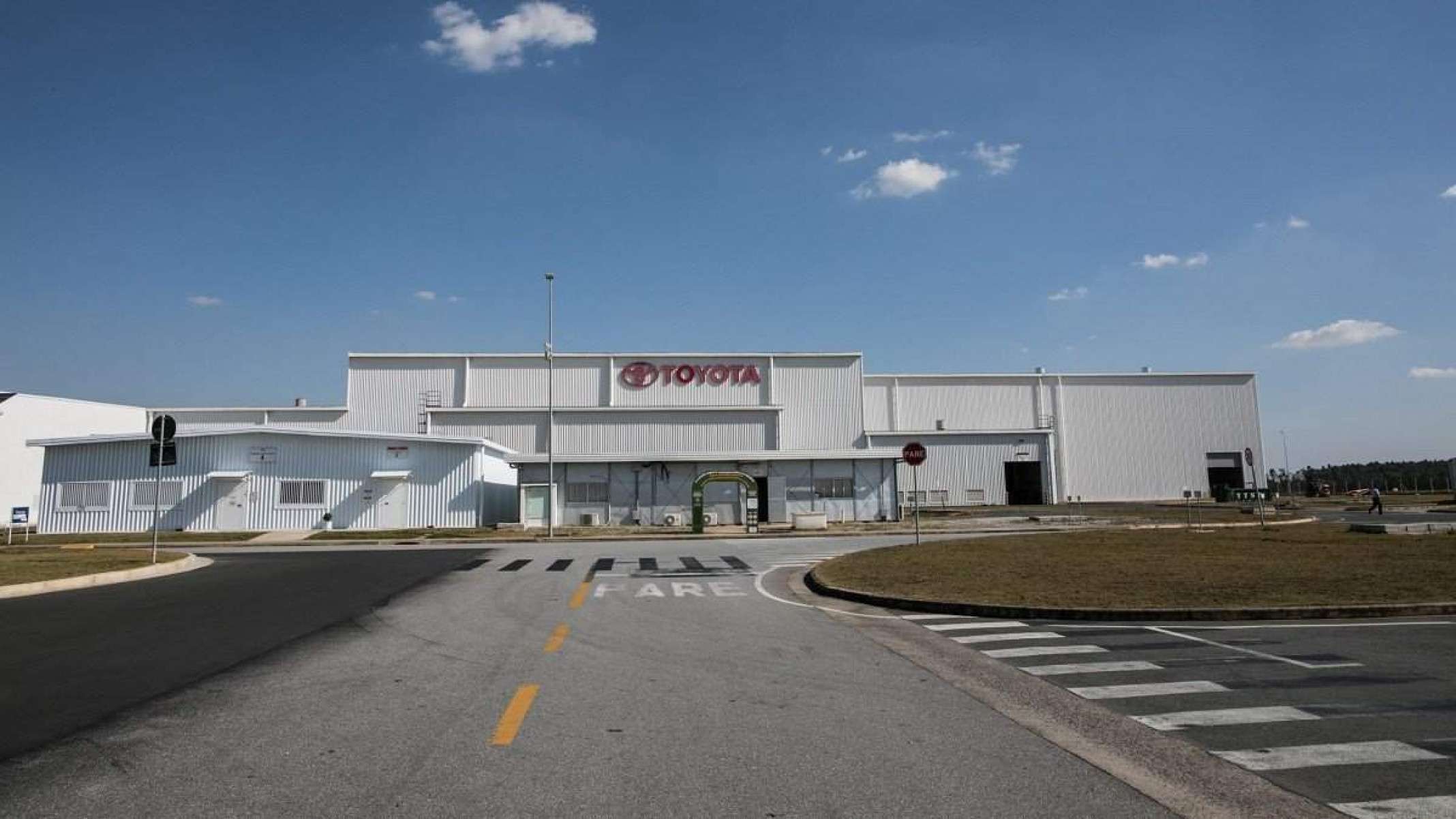 Imagem mostra entrada de fábrica da Toyota em Sorocaba (SP), prédio é branco e possui letreiro com nome da montadora em vermelho. Em primeiro plano é possível ver via de acesso para a planta e ao fundo céu claro com poucas nuvens