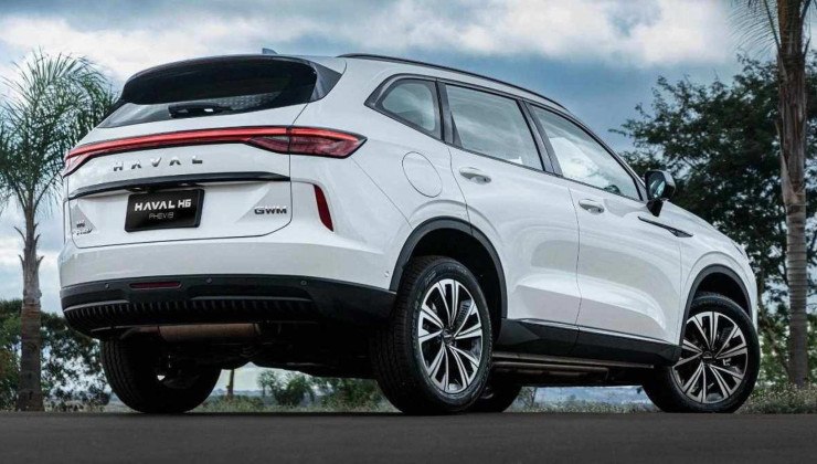 GWM Haval H6 PHEV19 branco visto pela traseira com detalhes da lateral em chão asfaltado. Ao fundo é possível ver palmeiras e céu claro com nuvens. 