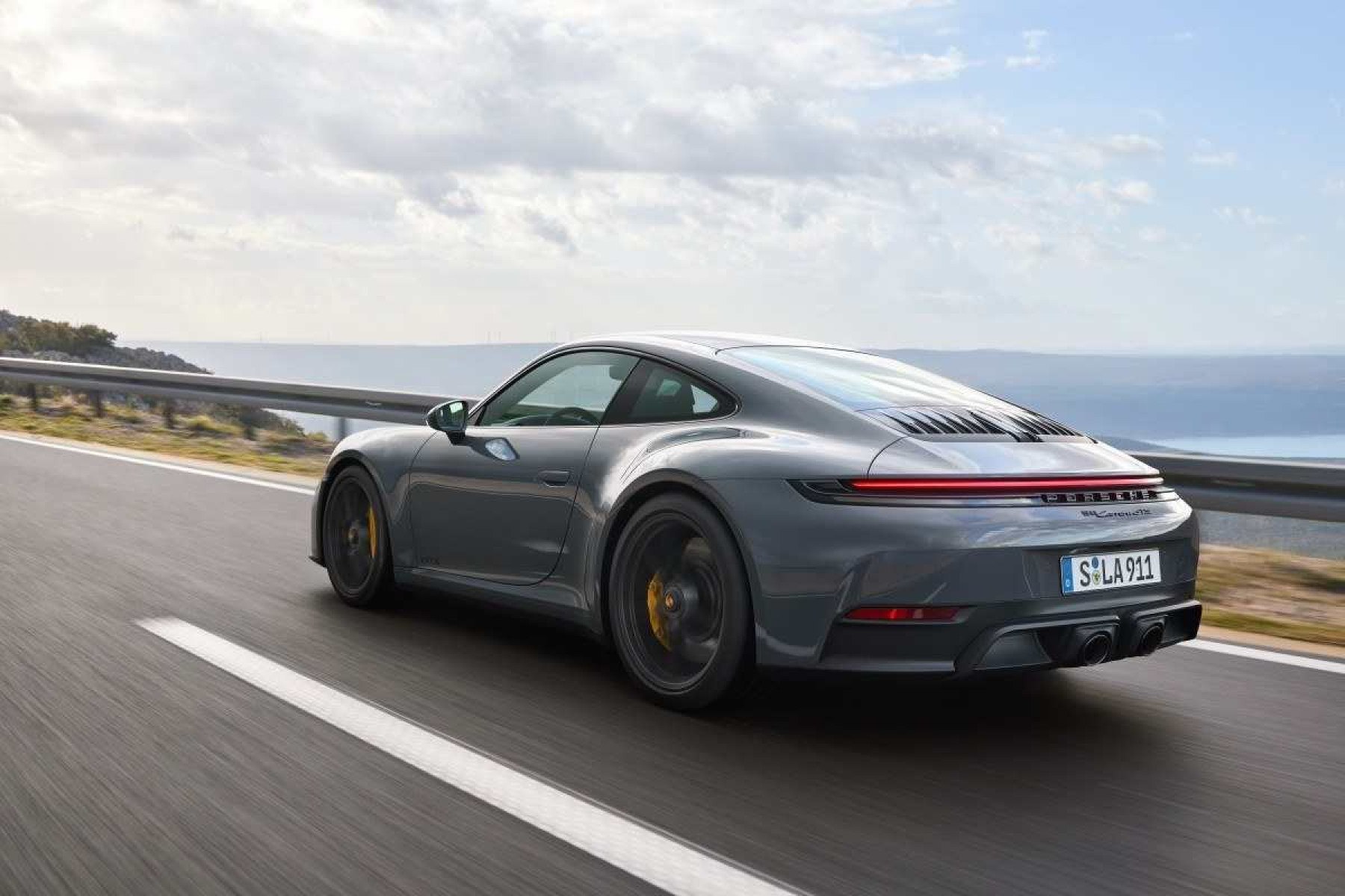 Porsche 911 Carrera GTS modelo 2024 cinza escuro de traseira em movimento na estrada asfaltada com mar ao fundo