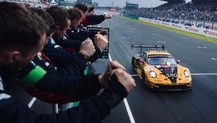 Porsche 911 GT3 R amarelo com detalhes em preto visto pela dianteira. Pessoas em arquibancada s&atilde;o vistas no canto esquerdo da imagem. Ve&iacute;culo est&aacute; em movimento em pista de corrida com faixas brancas na horizontal e vertical na via.
