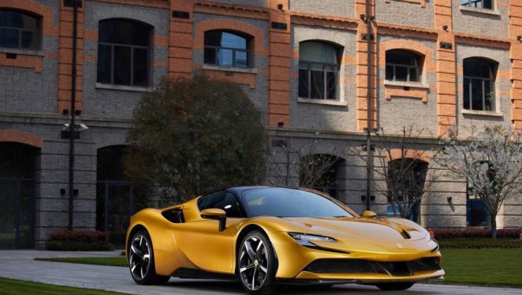 Ferrari SF90 Spider amarela, de frente, estacionada em uma estrada cercada de jardins, com um prédio histórico ao fundo