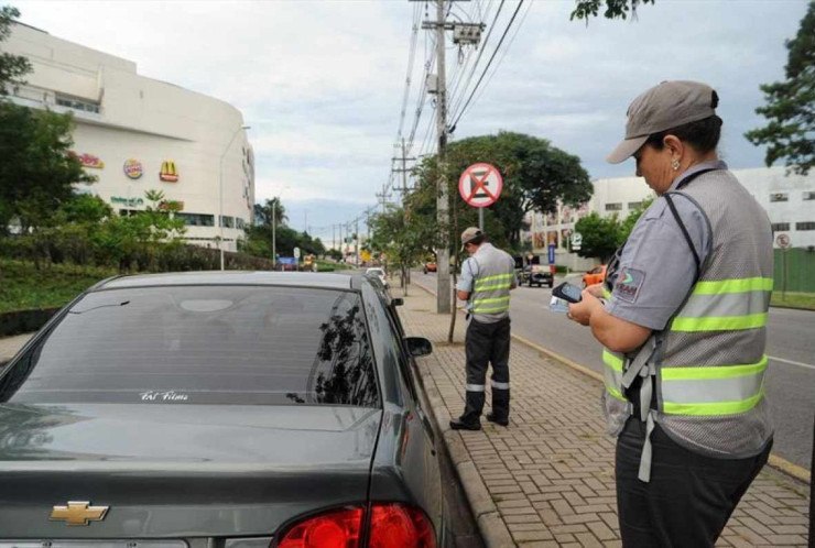 Guarda municipal de Curitiba com vestes cinzas e colete verde neon est&aacute; com bloco de multas na m&atilde;o. Pessoa est&aacute; parada ao lado de ve&iacute;culo prata, ao fundo &eacute; poss&iacute;vel ver outro guarda e c&eacute;u nublado. 
