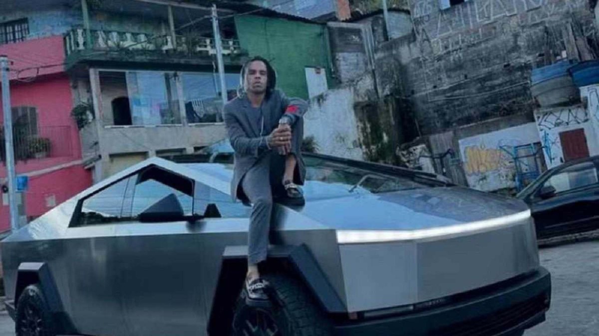 Danielzinho Grau de jaqueta, calça social e sapato preto está sentado no capô de um Cybetruck prateado.  Ao fundo avistam-se as ruas do Morro do Macaco.