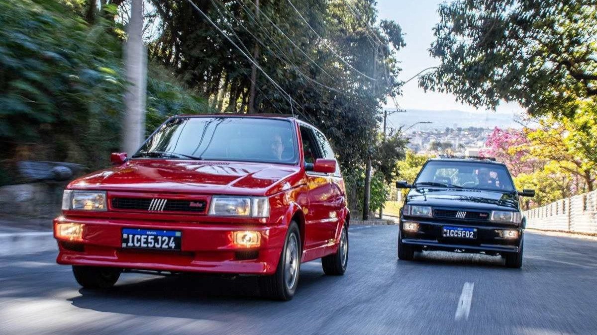 Duas unidades do Fiat Uno Turbo 1994, uma preta e a outra vermelha, de frente, em movimento, em avenida urbana