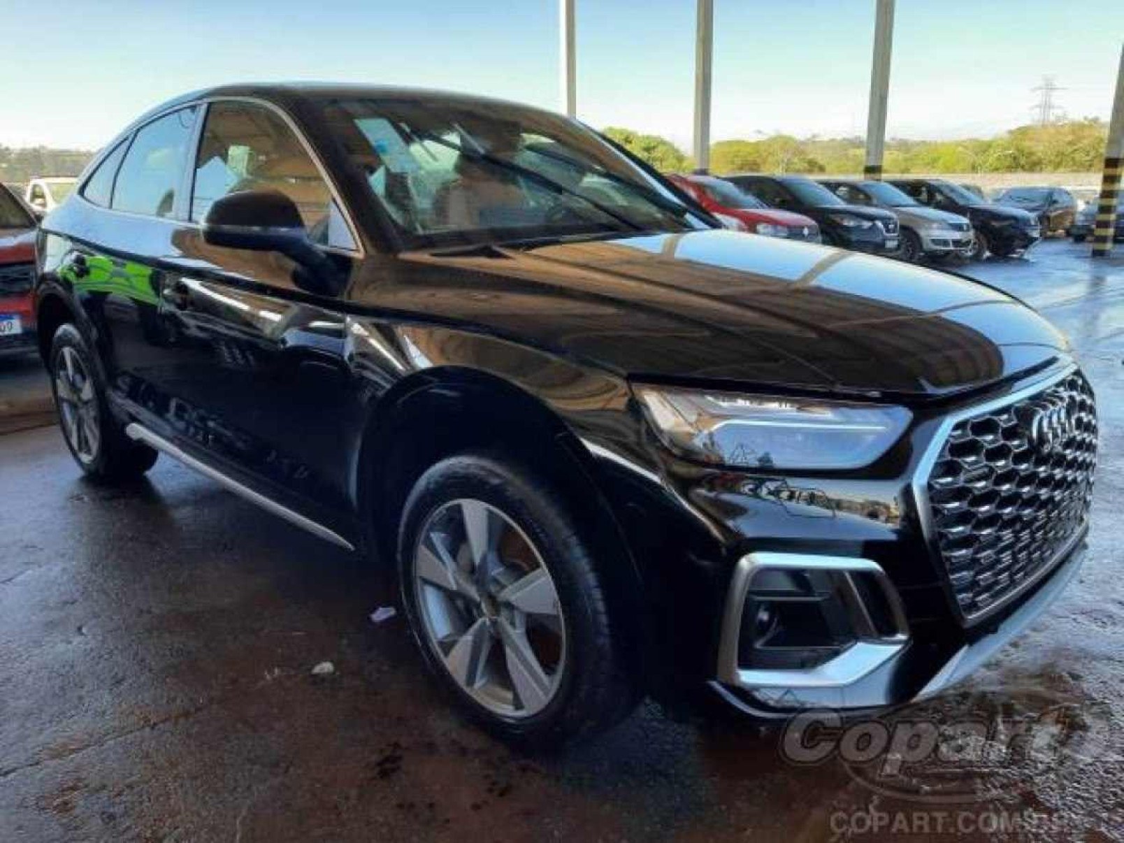 Audi Q5 preto visto pela dianteira com detalhes da lateral esquerda estacionado em chão de pátio. Céu claro é visto ao fundo