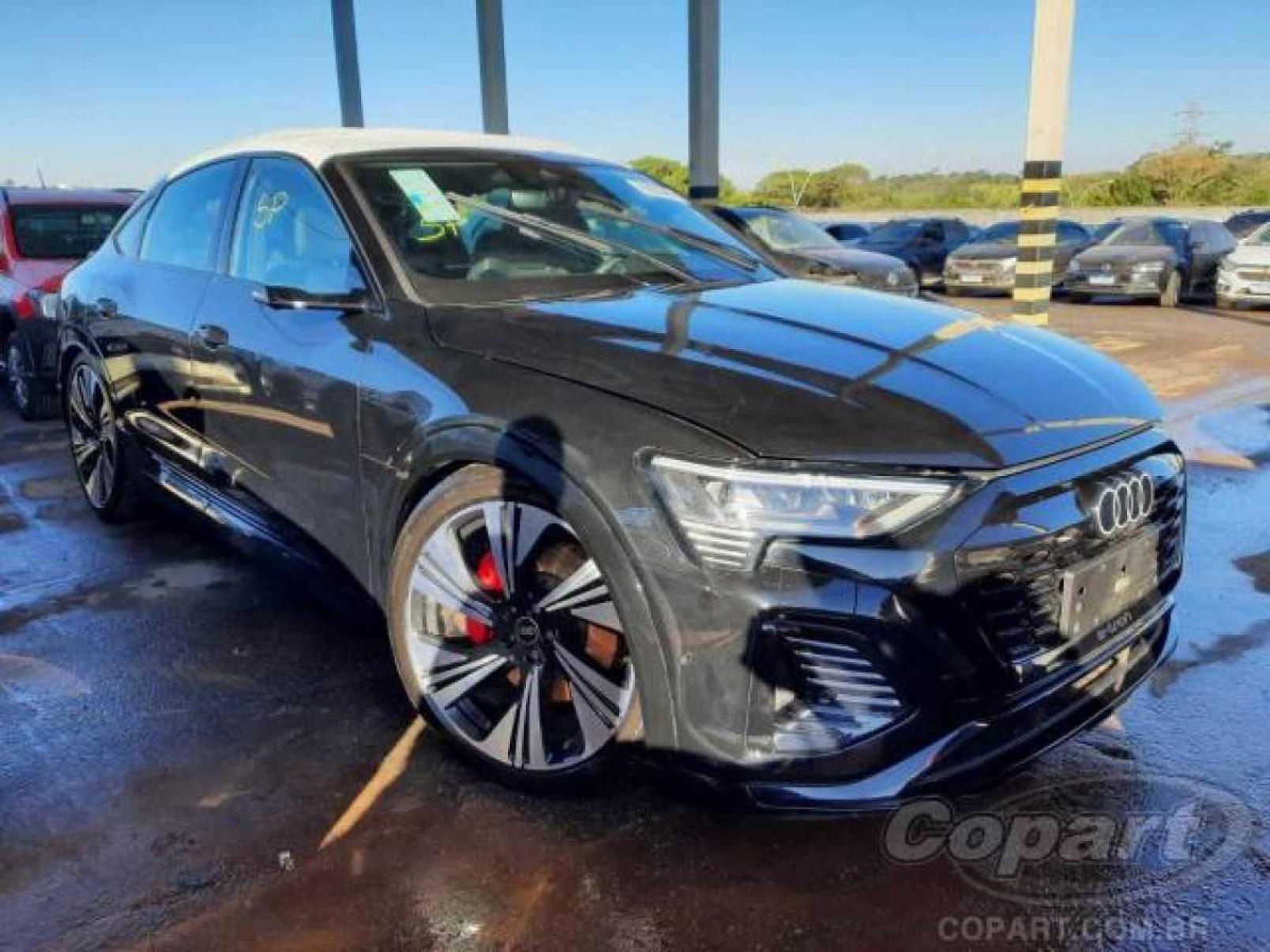 Audi Q8 cinza visto pela dianteira com detalhes da lateral esquerda estacionado em chão de pátio. Sombras batem na carroceria do carro, céu claro e arbustos são vistos ao fundo