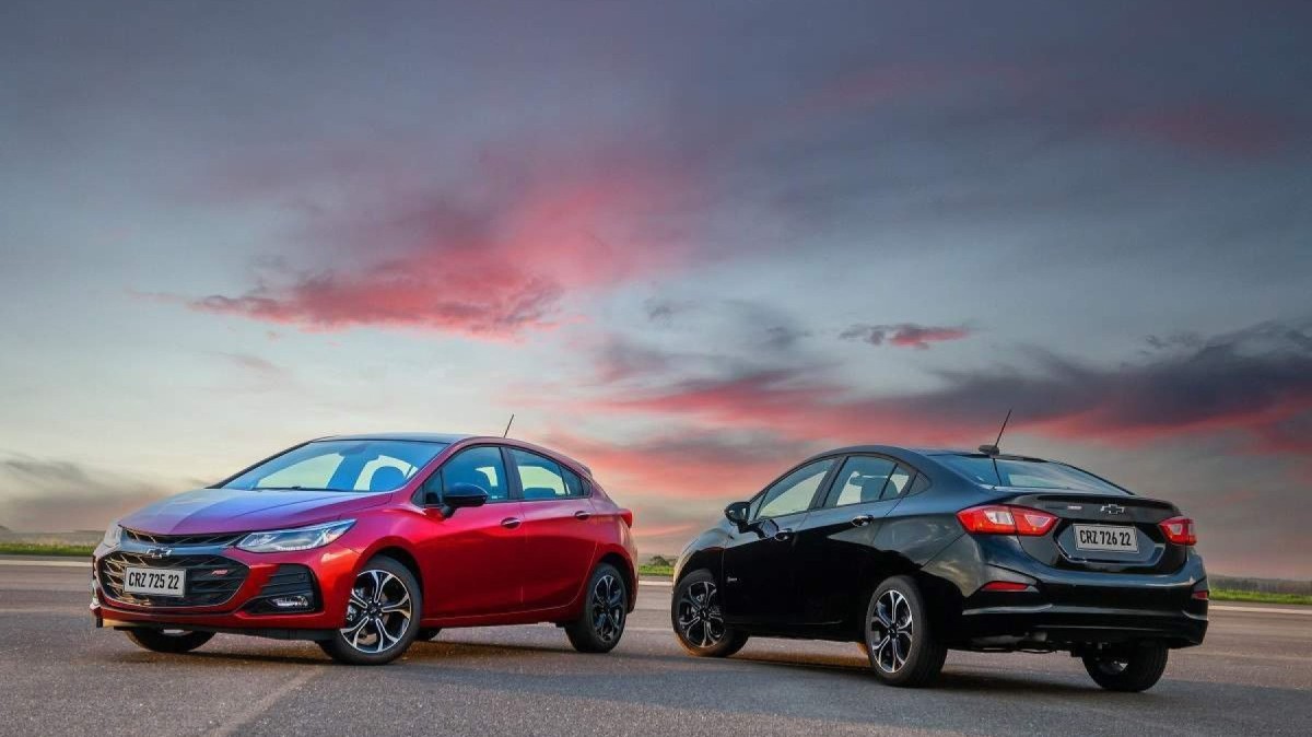 Chevrolet Cruze hatch RS vermelho, na frente, e Cruze sedã preto Midnight, na traseira, estacionados a 90 graus um do outro, em local asfaltado, com o pôr do sol ao fundo