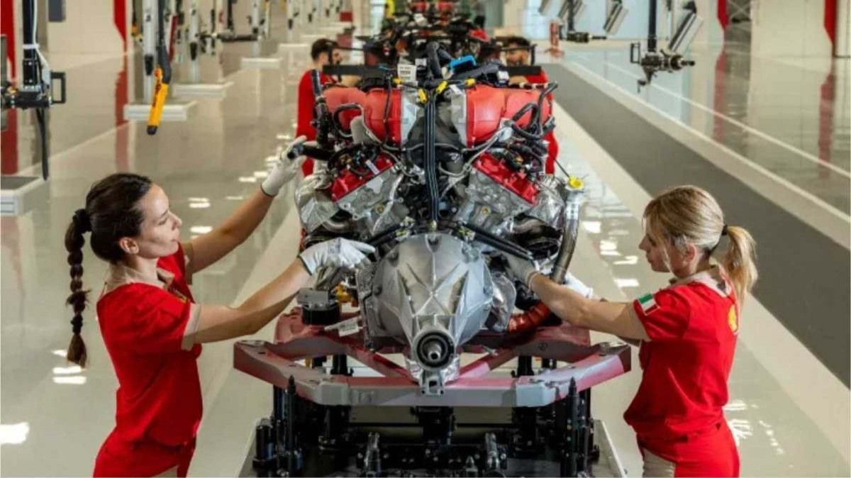 Interior da fábrica da marca Ferrari.  No meio, aparece um motor, em um dos postos da fábrica, sendo operado por duas mulheres.  À direita, uma mulher loira com uniforme vermelho, e à esquerda, uma mulher de cabelos pretos usando uniforme vermelho.