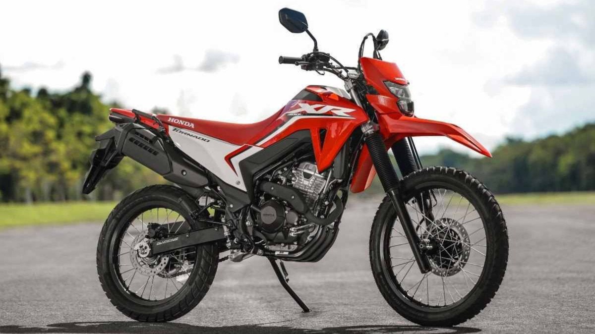 Modelo Honda XR 300L Tornado 2024 vermelho com detalhes laterais estáticos brancos no asfalto com árvores ao fundo