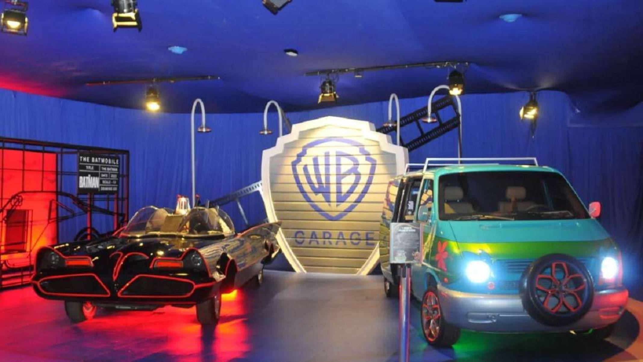 Batmóel e Máquina de Mistério parados de frente em exposição. Ambos os veículos são vistos pela dianteira e iluminados por luzes. Ao fundo é possível ver parede azul com porta que se assemelha ao logo da Warner Bros escrito "WB Garage"