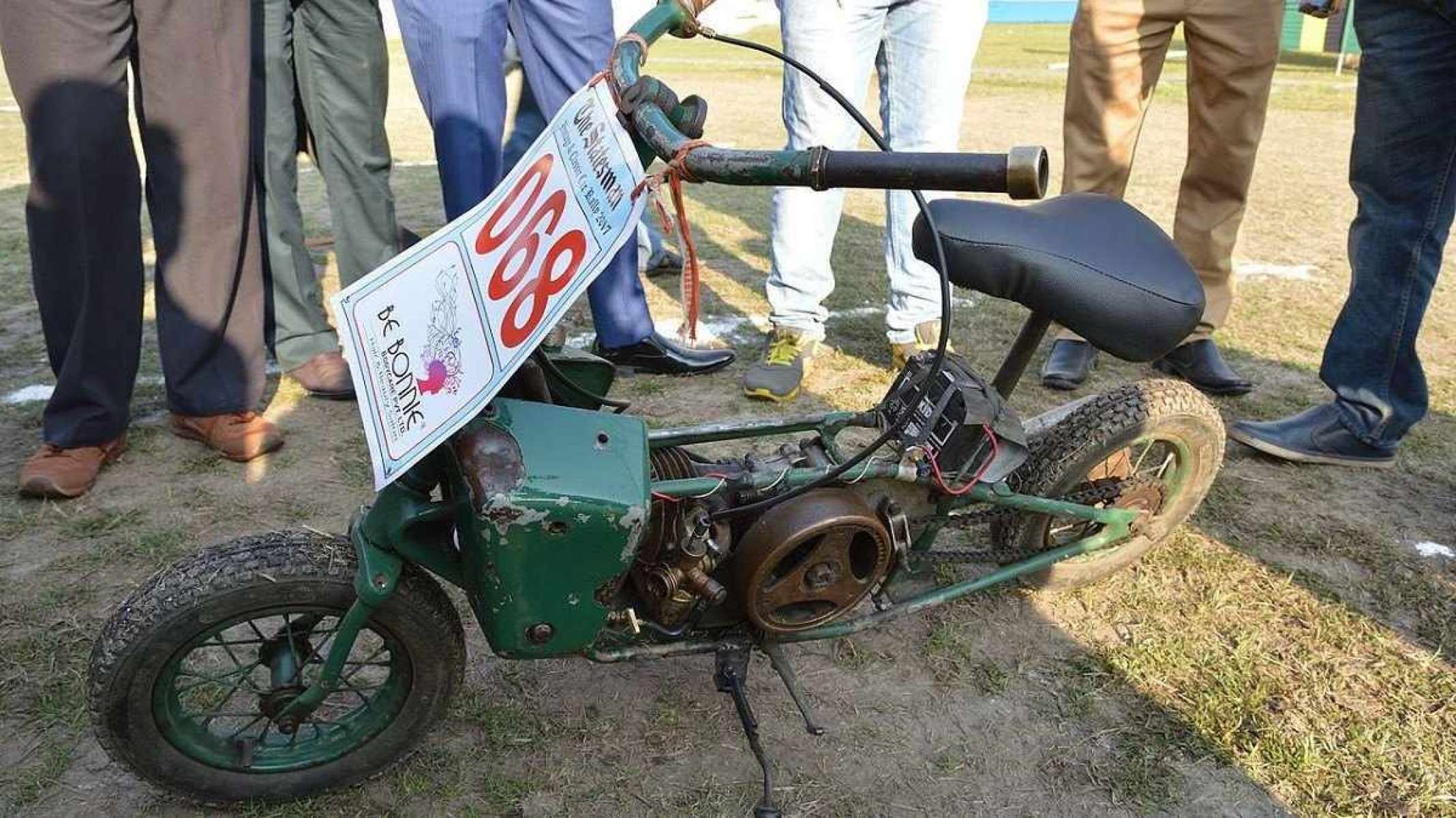 Welbike verde musgo estacionada é vista pela lateral esquerda. Ao fundo diversas pernas utilizando calças jeans claras e escuras são vistas em gramado