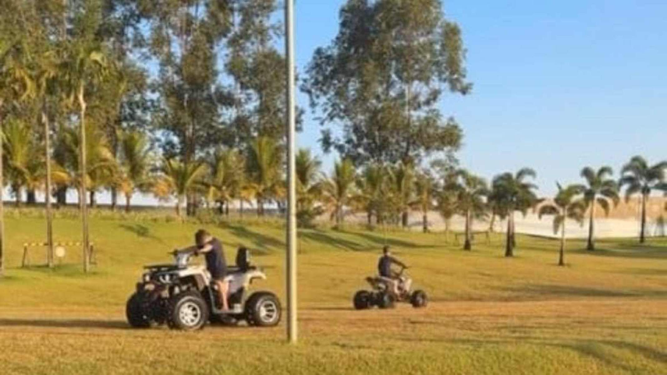 Foto mostra dois garotos brancos com blusas e bermudas pretas conduzindo quadriciclos. A imagem foi feita em gramado com árvores estreitas ao fundo e céu claro sem nuvens acima