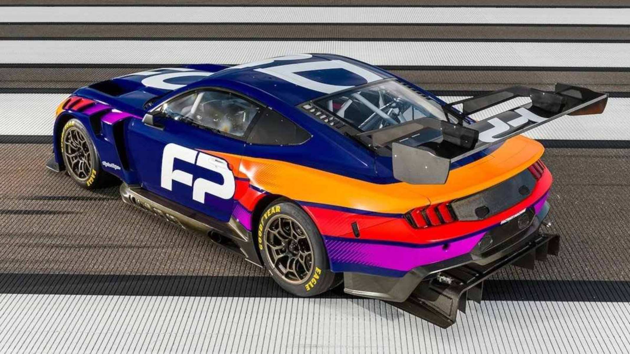 Ford Mustang GT3 nas cores azul, laranja, vermelho e roxo parado de traseira em pista de corrida visto de cima 