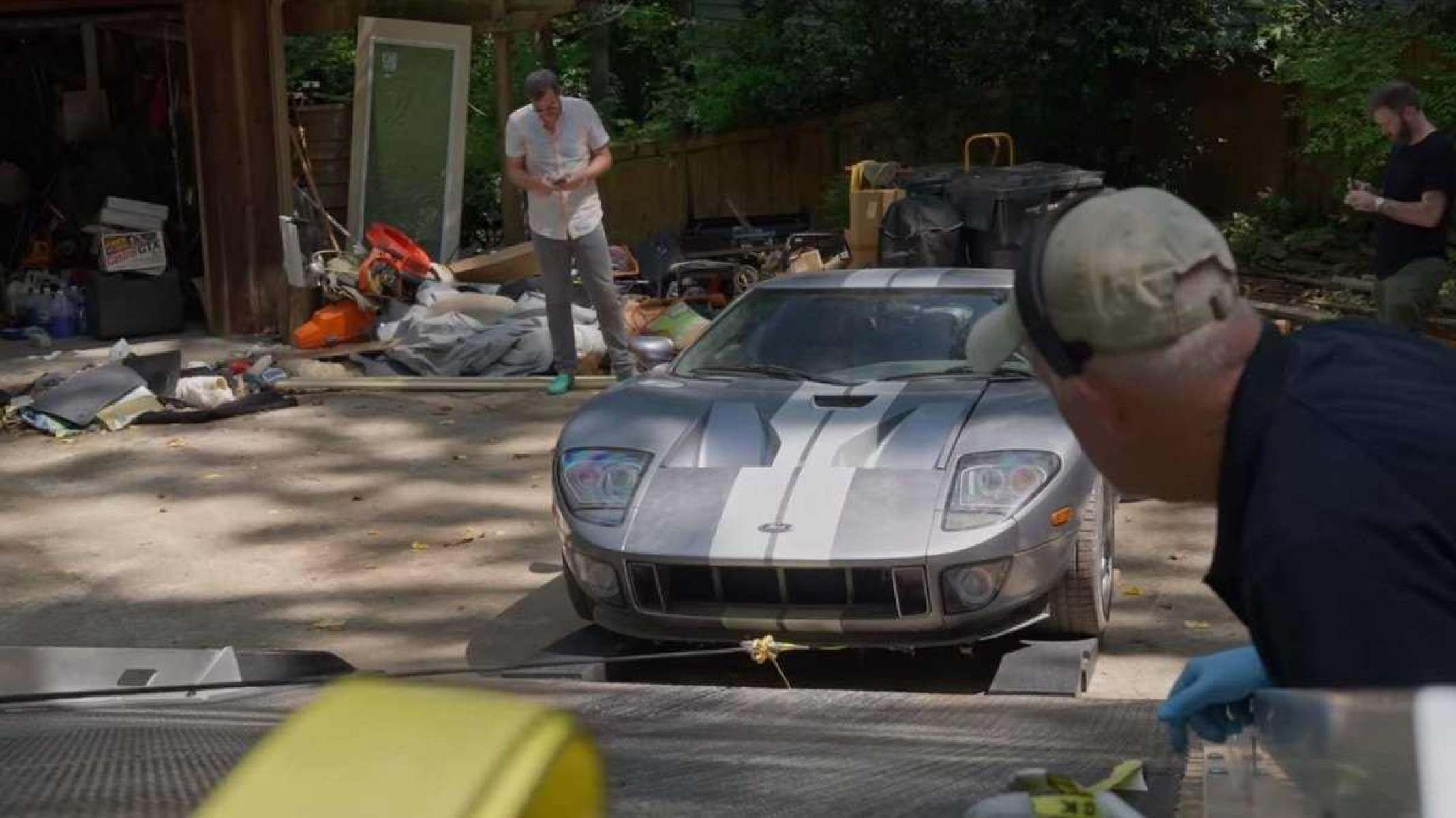 O Ford GT 2006 cinza frontal está sendo puxado para a rampa de reboque.  Um homem de boné cinza está em primeiro plano, enquanto algumas pessoas observam a cena ao fundo.  É possível ver uma casa borrada atrás