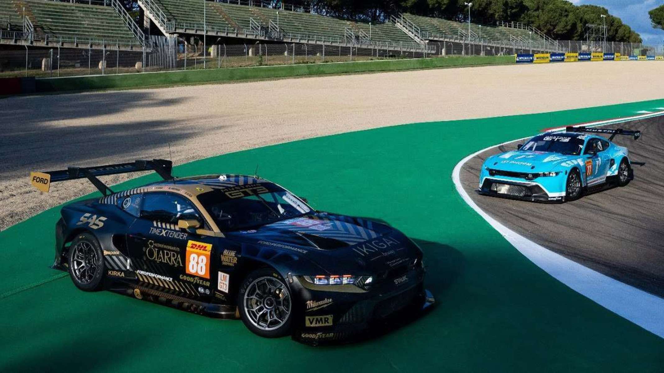 Dois Ford Mustang GT3 um preto e outro azul, ambos de frente, parados em pista de corrida