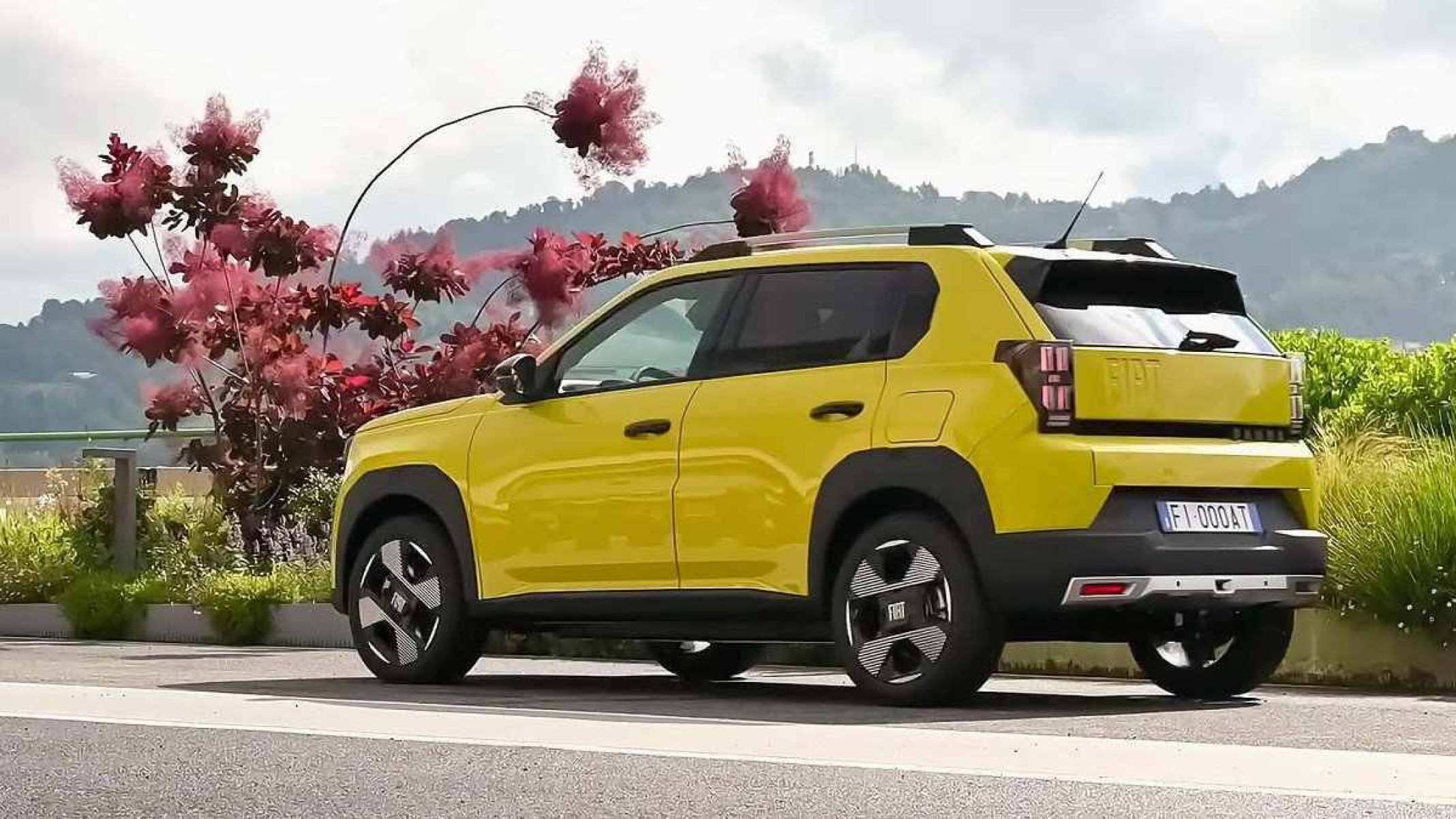 Fiat Grande Panda amarelo visto do lado esquerdo e traseiro.  Veículo trafega em estrada pavimentada.  Ao fundo você pode ver arbustos e plantas com folhagem vermelha.  Céu coberto por nuvens e montanha coberta por vegetação ao fundo