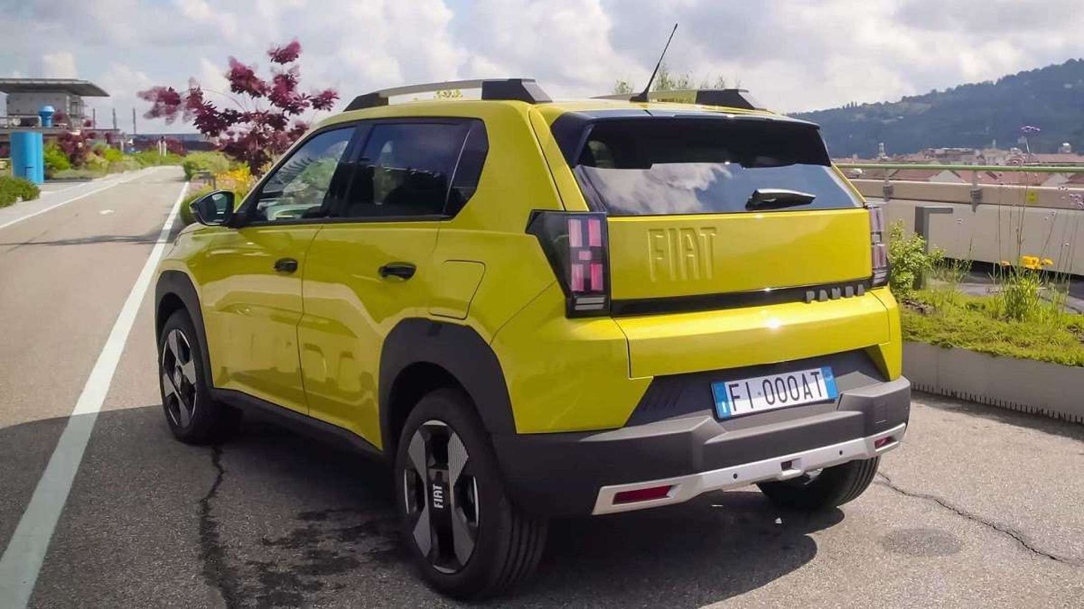 Fiat Grande Panda amarelo visto de trás e do lado esquerdo.  Veículo está parado em uma estrada de asfalto com linhas verticais brancas.  Ao fundo é possível observar vegetação com folhagens vermelhas e um céu claro coberto por nuvens.