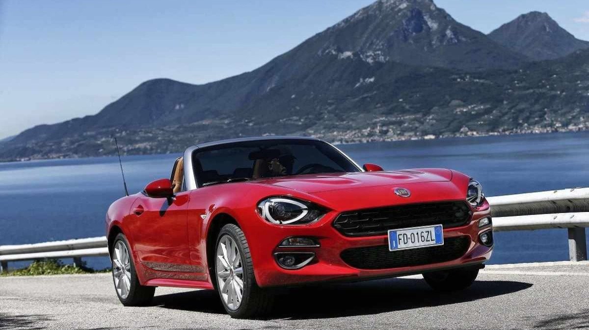 Red Fiat 124 Spider, movendo-se na estrada, com lago e montanhas ao fundo