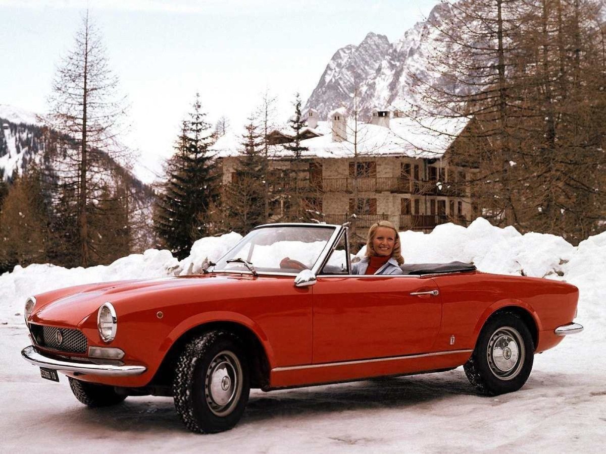 Fiat 124 Sport vermelho, estacionado em local nevado, com a capota aberta e mulher ao volante