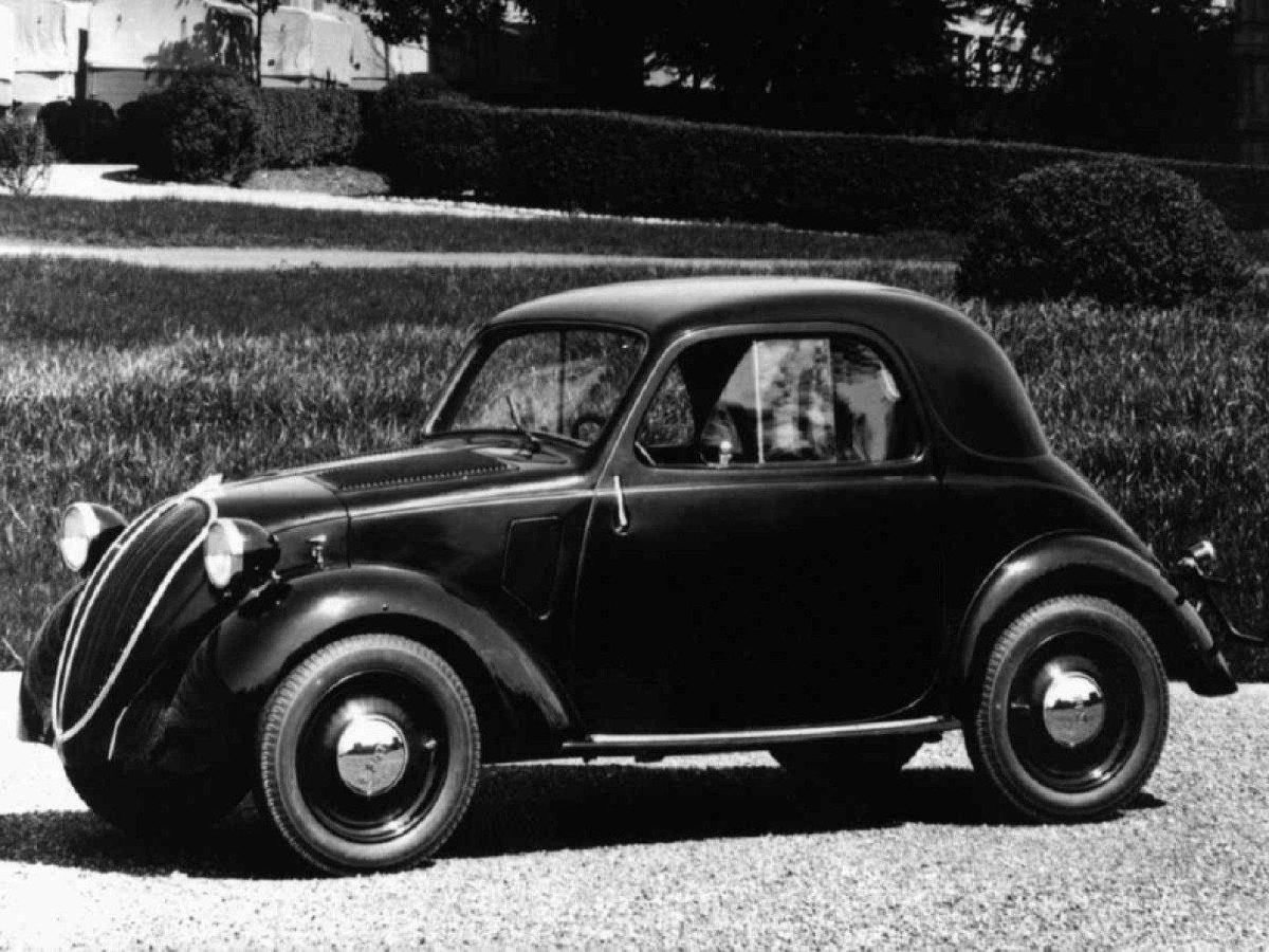 Fiat 500 Topolino, preto, de lado, estacionado, com jardim ao fundo, em foto preto e branca